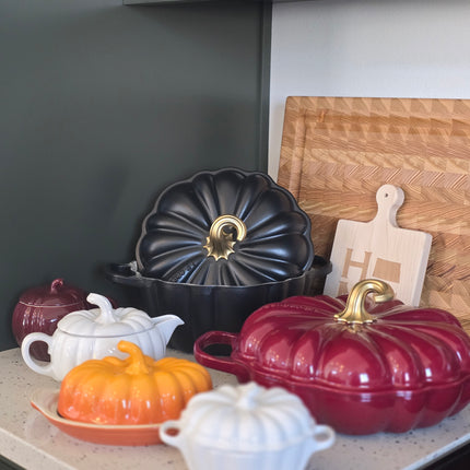 a collection of colorful, decorative kitchenware, including a red and black pot, a black pot with a gold handle, and several white and orange pots, all arranged on a kitchen counter.