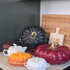 a collection of colorful, decorative kitchenware, including a red and black pot, a black pot with a gold handle, and several white and orange pots, all arranged on a kitchen counter.