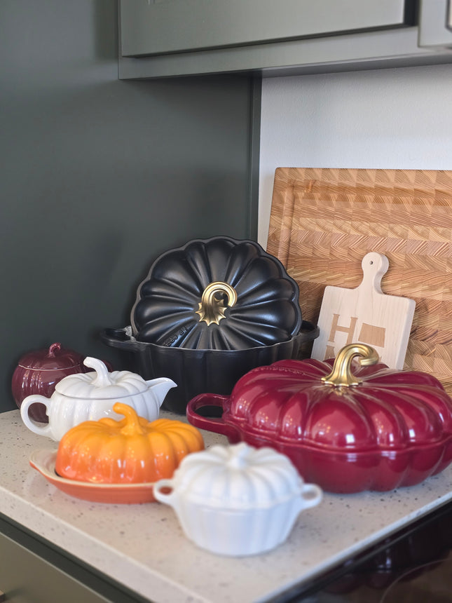a collection of colorful, decorative kitchenware, including a red and black pot, a black pot with a gold handle, and several white and orange pots, all arranged on a kitchen counter.