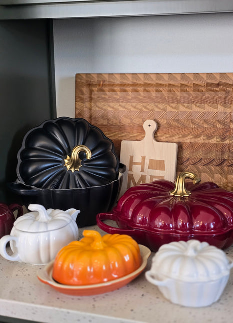 a collection of kitchen utensils and containers, including a wooden cutting board, a black pot lid, a red pot lid, a white teapot, and a wooden tray.