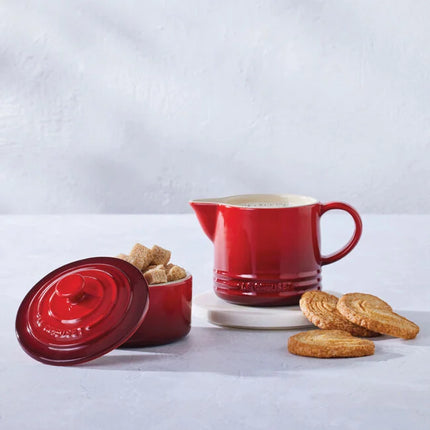 a red ceramic pitcher, a red ceramic container with a lid, and a plate of cookies on a white surface.