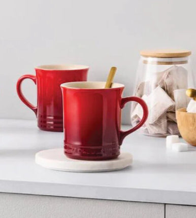 two red mugs with handles, one of which has a spoon inside, placed on a white surface. There is also a glass jar with a lid and a wooden bowl with a spoon inside.